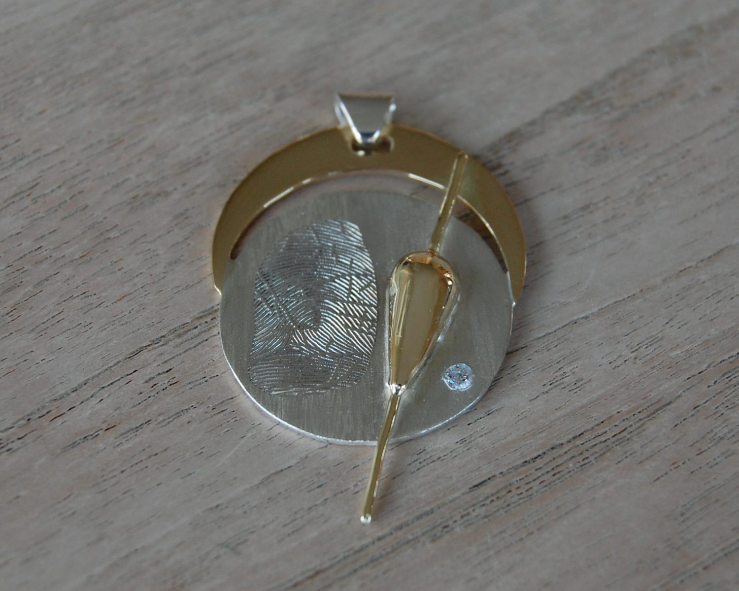 Symbolische hanger met een basis in zilver en accenten uit deels aangeleverd goud. In de dobber zit as en er is een vingerafdruk in het zilver gelaserd - inclusief zirkonia.